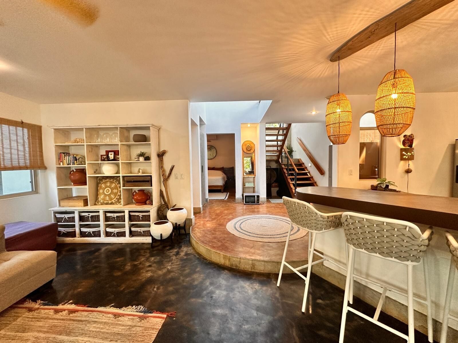 A modern living room with a dark floor, a white sofa, a coffee table, and a bookshelf. There are two wicker lamps hanging from the ceiling.