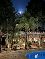Un área de piscina al aire libre con un espacio de descanso cubierto, palmeras e iluminación ambiental.