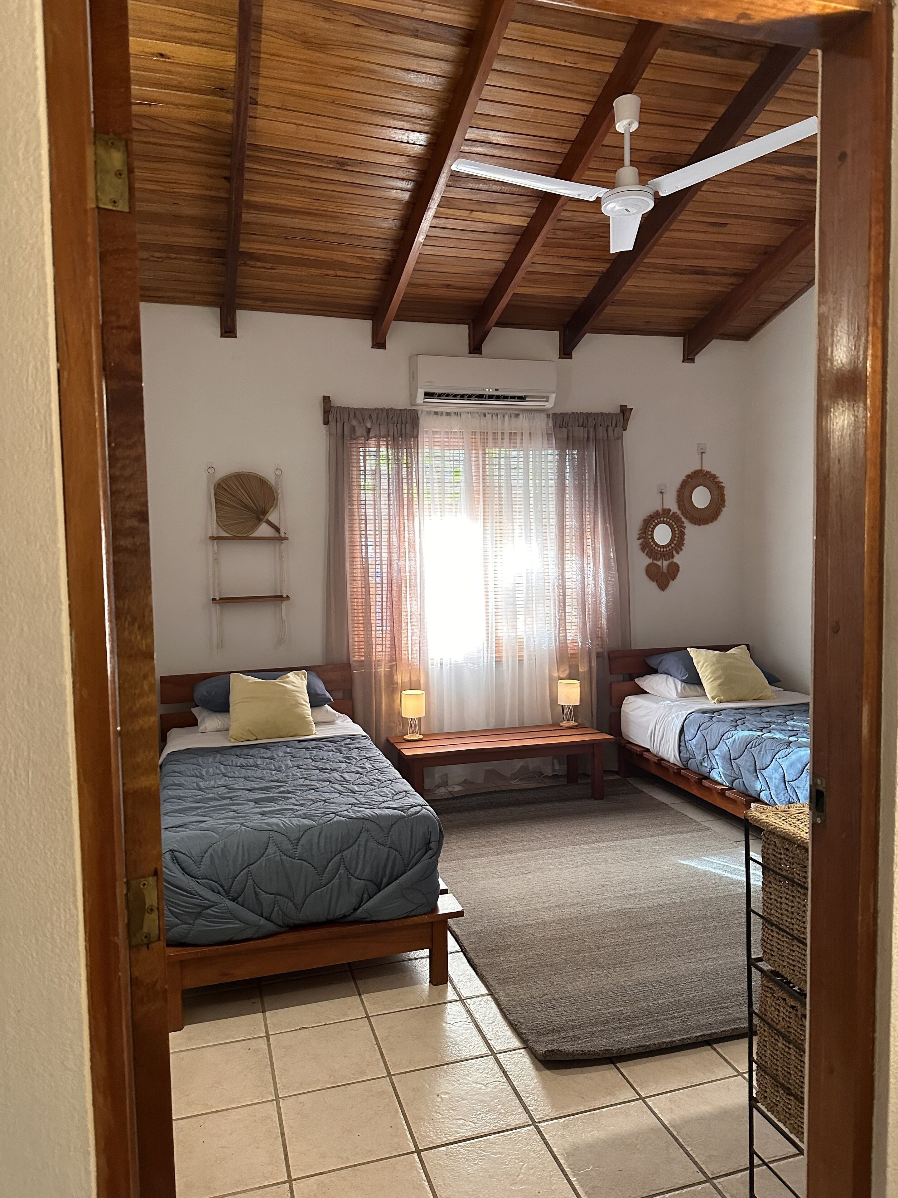 Un dormitorio con dos camas, ventilador de techo, equipo de aire acondicionado y una mesita con lámparas.
