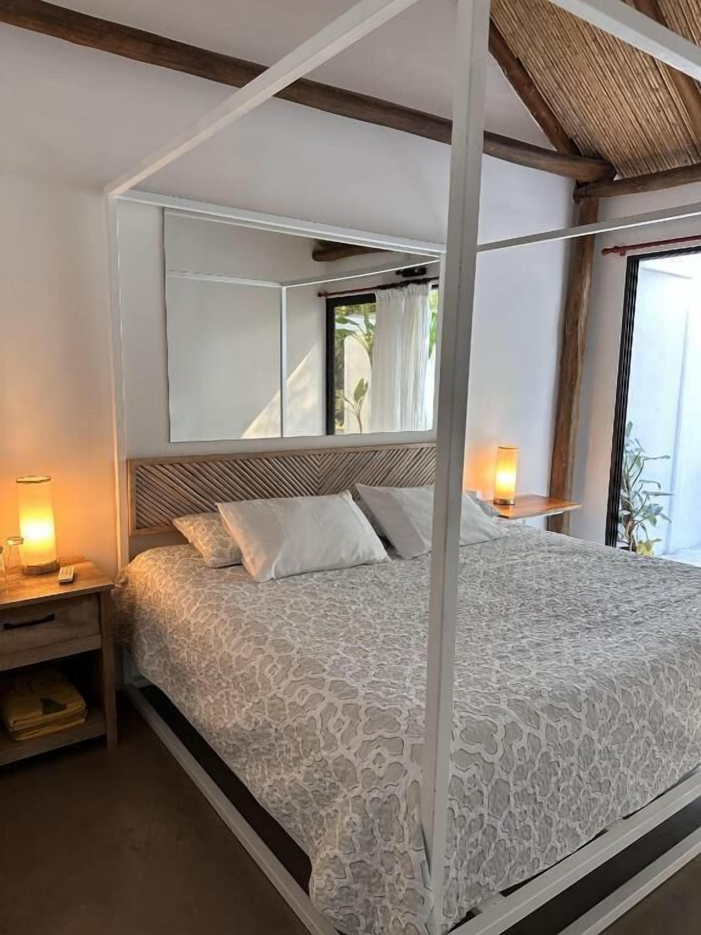 A four-poster bed with a patterned bedspread, bedside lamps, and a view of greenery outside.