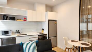 Cocina moderna con armarios blancos, un refrigerador de acero inoxidable, un microondas y una cafetera. Hay un sofá negro con un cojín azul y blanco, y una pequeña mesa de comedor con dos sillas blancas.