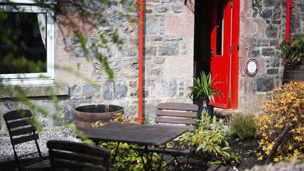 A stone building with a red door, outdoor seating, and a window.