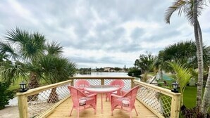 A wooden deck with a table and chairs, surrounded by a fence and palm trees. - Cozy Coastal Retreat, Steps to the Sugar Sand Beach of FMB! (Fort Myers Beach)