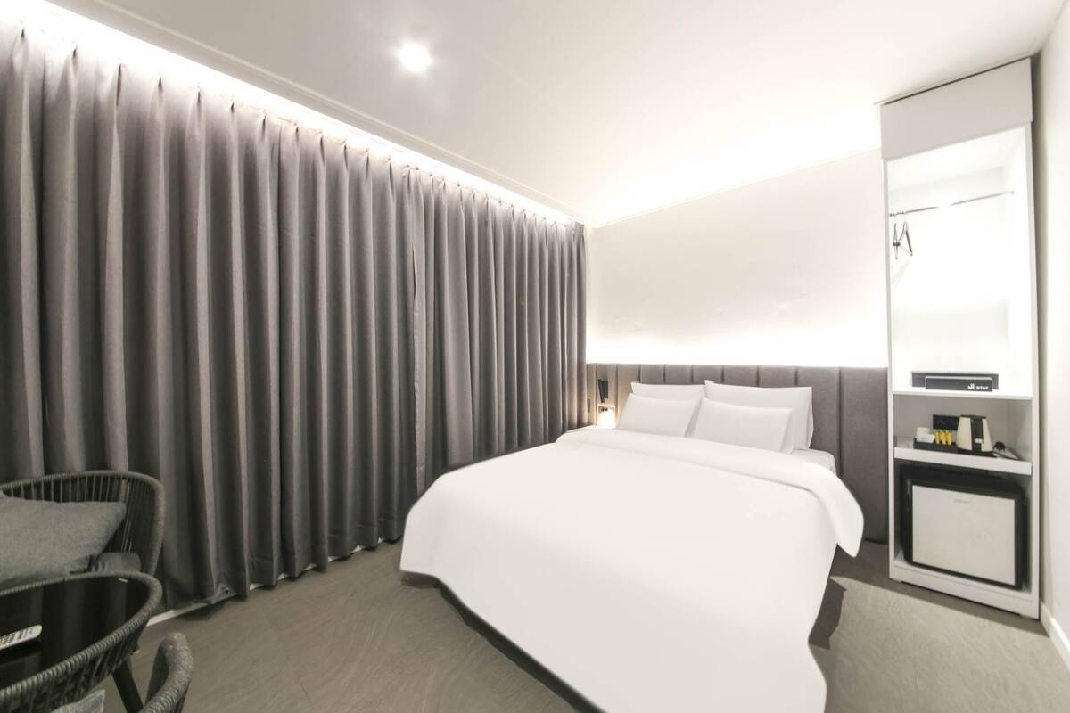 Ein modernes Hotelzimmer mit einem großen Bett, grauen Vorhängen, einer kleinen Sitzecke und einem Spiegel.