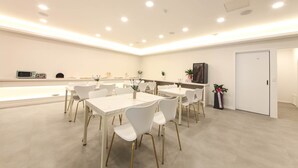 A spacious, well-lit room with multiple white tables and chairs, a kitchen area with appliances, and a door on the right. - JI STAY Hotel Gyeongju (Gyeongju)