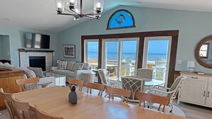 A spacious living area with a large wooden dining table, comfortable seating, a fireplace, and a view of the ocean through large windows. - Edgewater - Oceanfront with panoramic views! 4 bedroom, 3 baths, dog friendly and spa bath! (Southern Shores)