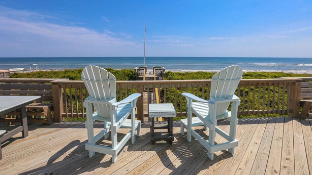 A wooden deck with two Adirondack chairs and a small table, overlooking a beach with a wooden fence and a clear sky. - Edgewater - Oceanfront with panoramic views! 4 bedroom, 3 baths, dog friendly and spa bath! (Southern Shores)