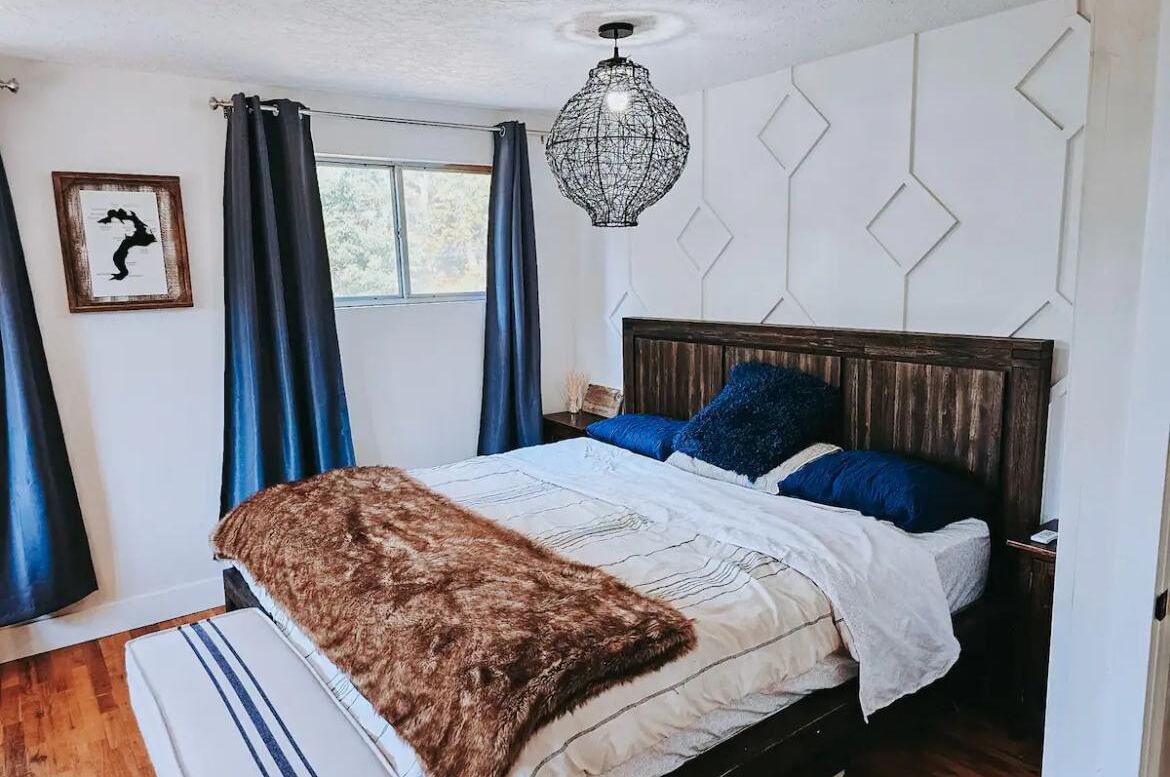 A bedroom with a wooden headboard, a large window with blue curtains, a hanging light fixture, and a framed picture on the wall.
