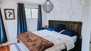 A bedroom with a wooden headboard, a large window with blue curtains, a hanging light fixture, and a framed picture on the wall.