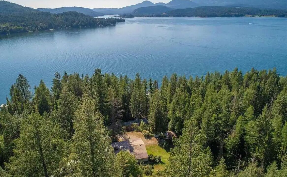 Ilmakuva metsäisestä alueesta, jossa on järvi ja pieni talo, jonka ympärillä kasvaa puita.