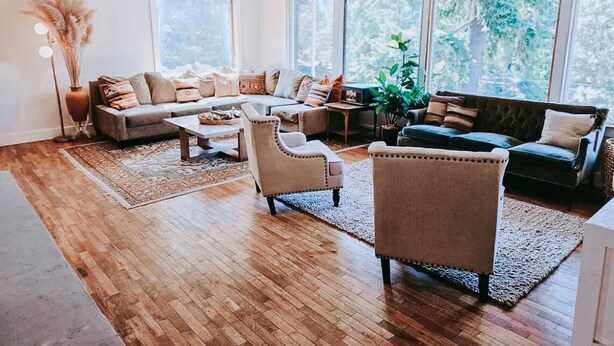 Un salon spacieux doté d’un plancher en bois, d’un canapé d’angle, d’une table basse et d’une grande fenêtre donnant sur la verdure.