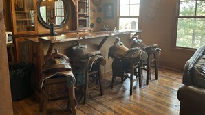 A rustic interior with wooden furniture, a bar area, and a large window with a view of trees. - Rustic 4 bedroom cabin at Gilded Mountain, wifi, pool access, paved roads (Lead)