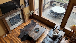 A cozy living room with a stone fireplace, leather sofa, and a large window overlooking an outdoor deck. - Rustic 4 bedroom cabin at Gilded Mountain, wifi, pool access, paved roads (Lead)
