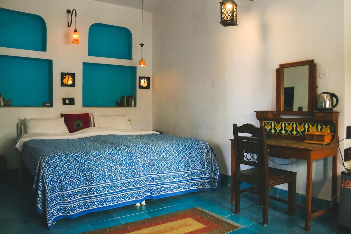 A bedroom with a bed, a desk, and a chair. There are wall-mounted shelves and a mirror.