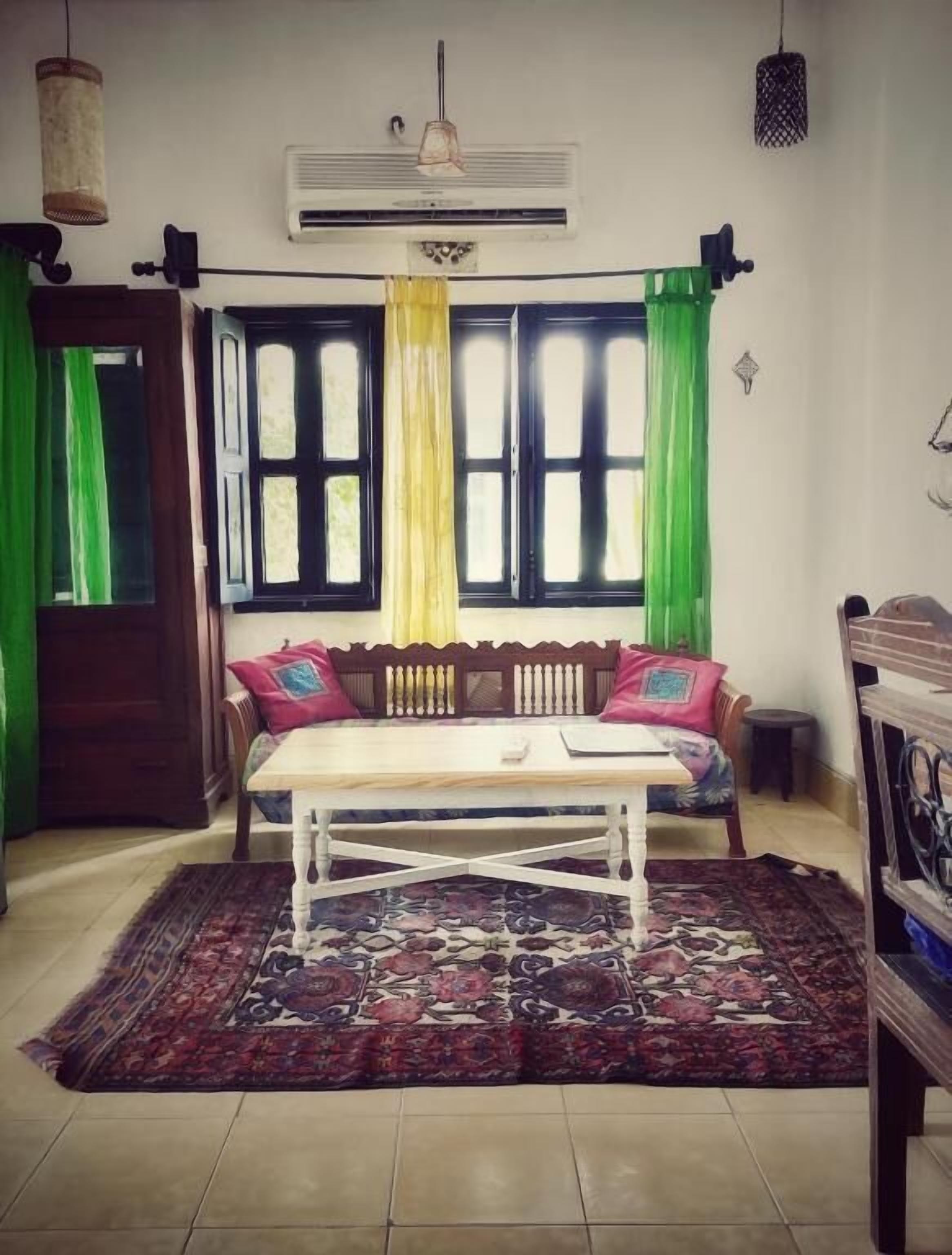 Una habitación con una mesa de madera, un tapete estampado, un sofá con cojines y una ventana con cortinas.