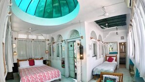 Une chambre avec deux lits, un ventilateur de plafond, une petite table avec un ordinateur portable, et une porte donnant sur une autre chambre.