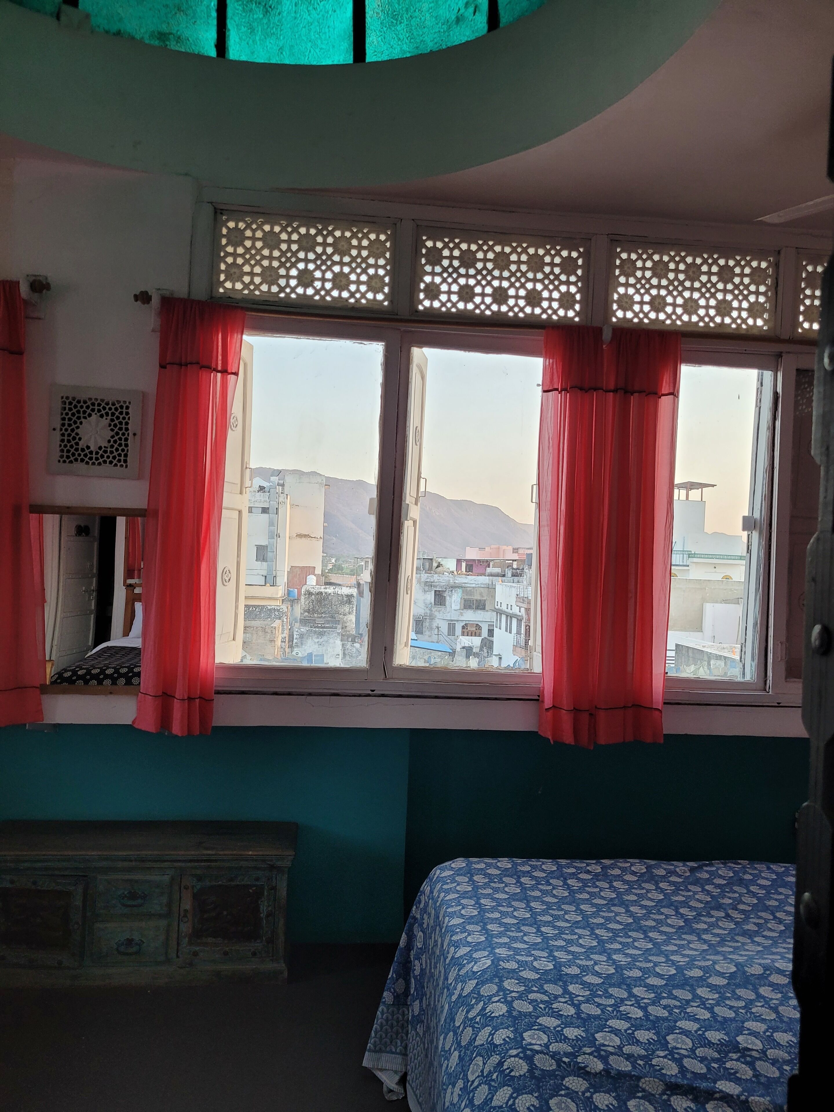 Une chambre avec un lit, une fenêtre ornée de rideaux rouges et une vue sur des immeubles et des montagnes.