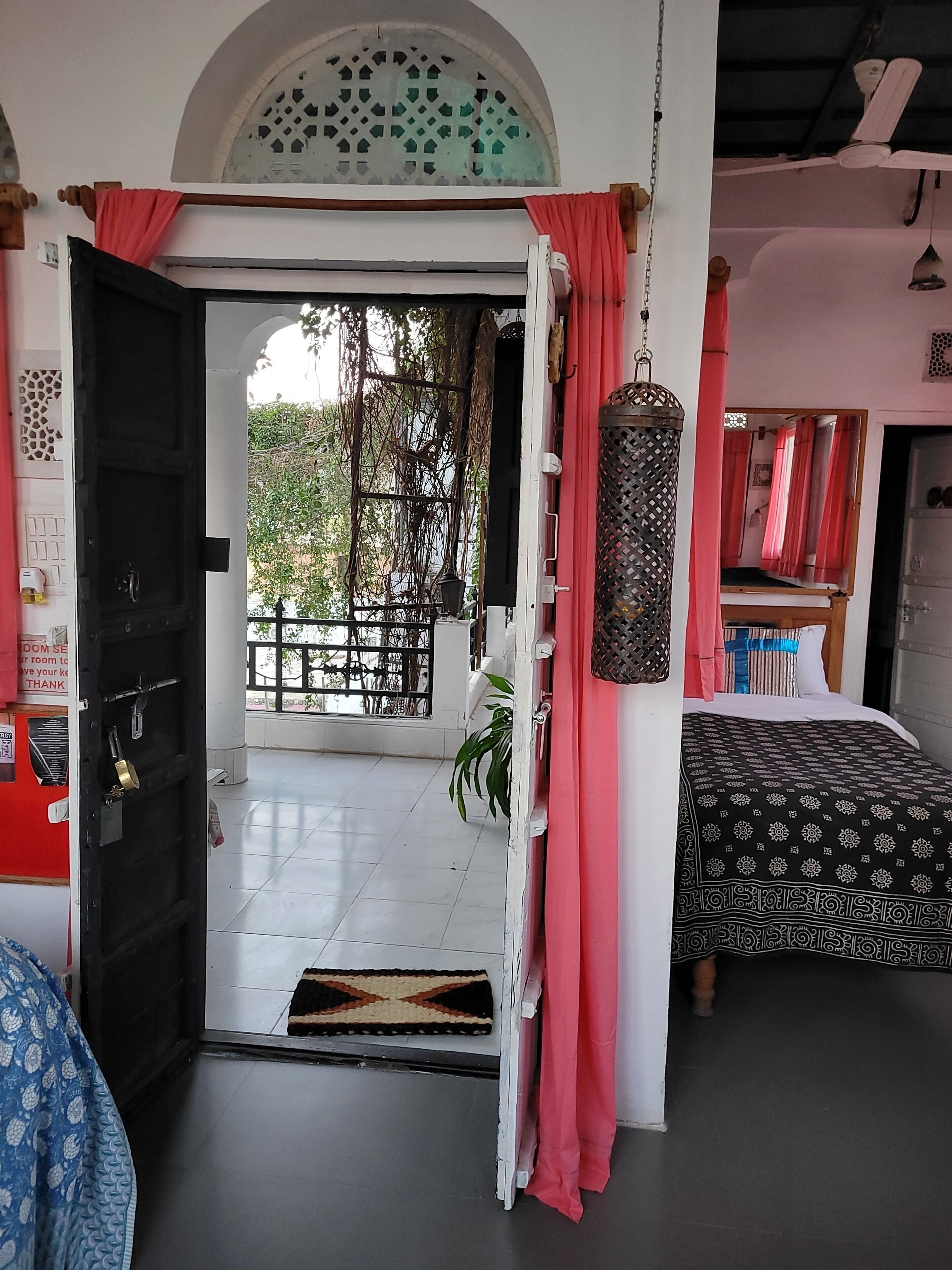 Una habitación con puerta negra, cama con colcha estampada, un farol colgante y vistas al verdor exterior.