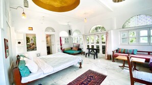 A spacious room with a large bed, a wooden bench, a small table, and a sofa. There are multiple windows with decorative glass panes. - Dia homestay (Pushkar)