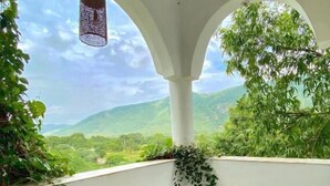 A balcony with two wicker chairs, a hanging light fixture, and a view of green hills. - Dia homestay (Pushkar)