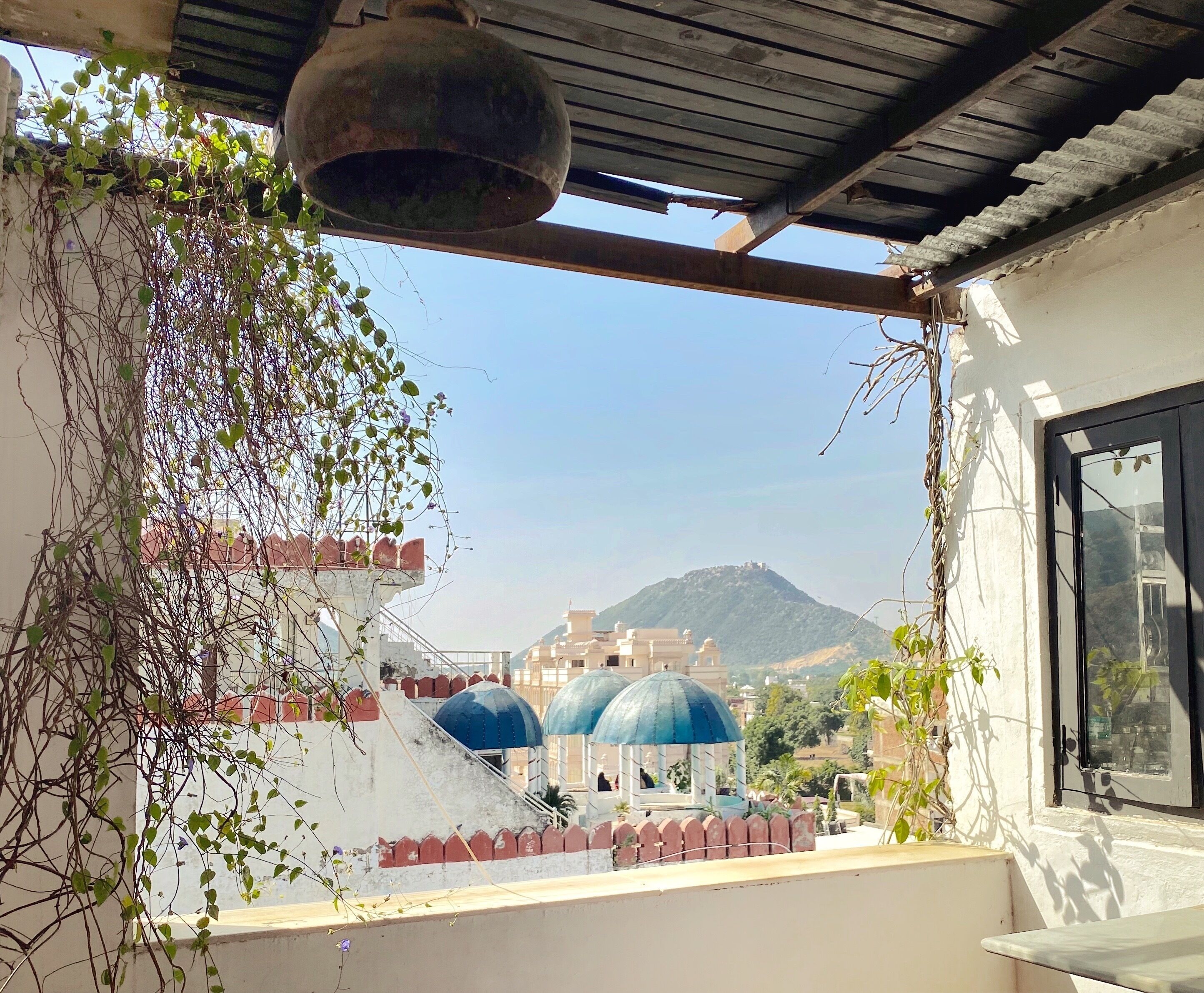 A balcony with a view of a mosque with blue domes, a mountain in the background, and a bell hanging above.