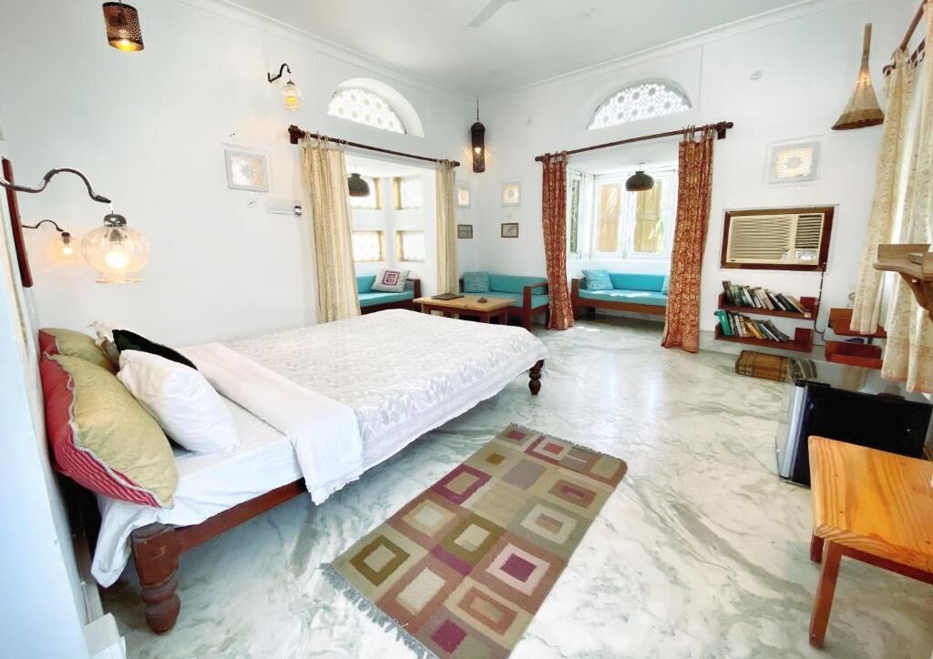 A room with a bed, a sofa, a small table, and a bookshelf. There are curtains, a lamp, and a window.