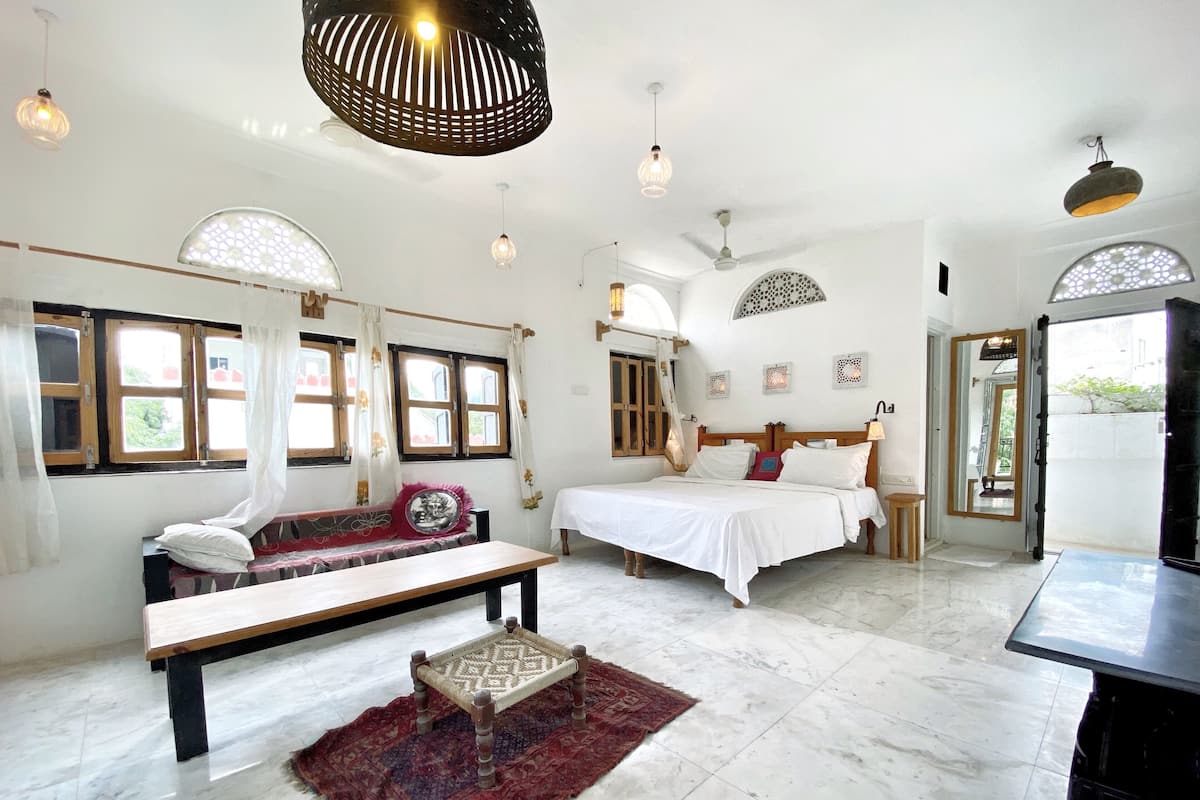 Una habitación amplia con una cama grande, un banco y una mesita. Varias ventanas ofrecen luz natural, y la habitación tiene un diseño moderno con paredes blancas y suelo de mármol.