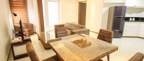 A modern living room with a glass-top dining table, brown leather sofas, and a kitchen area with white cabinets and a refrigerator.