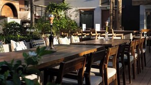 Un espacio al aire libre para comer con mesas y sillas de madera, rodeado de plantas en macetas y elementos arquitectónicos.