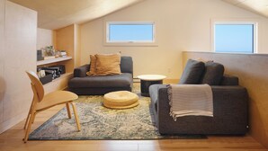 Een loft-achtige kamer met een hoge plafond, twee ramen, een bank, een stoel, een kleine ronde tafel en een boekenplank.