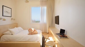 Een slaapkamer met een bed, een bureau met een laptop, een stoel, een televisie en een raam met gordijnen.