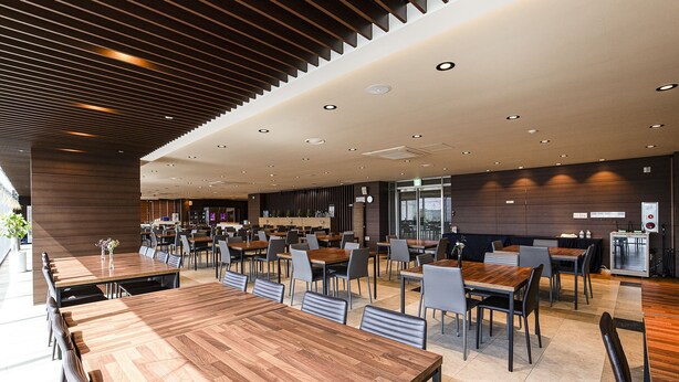 Uma ampla área de refeições com várias mesas e cadeiras de madeira, design moderno no teto e uma grande janela que proporciona luz natural.