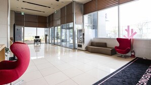 Um hall moderno com entrada por porta de vidro, máquina de vendas automática, uma cadeira vermelha, um sofá e uma janela grande com vista para o exterior.