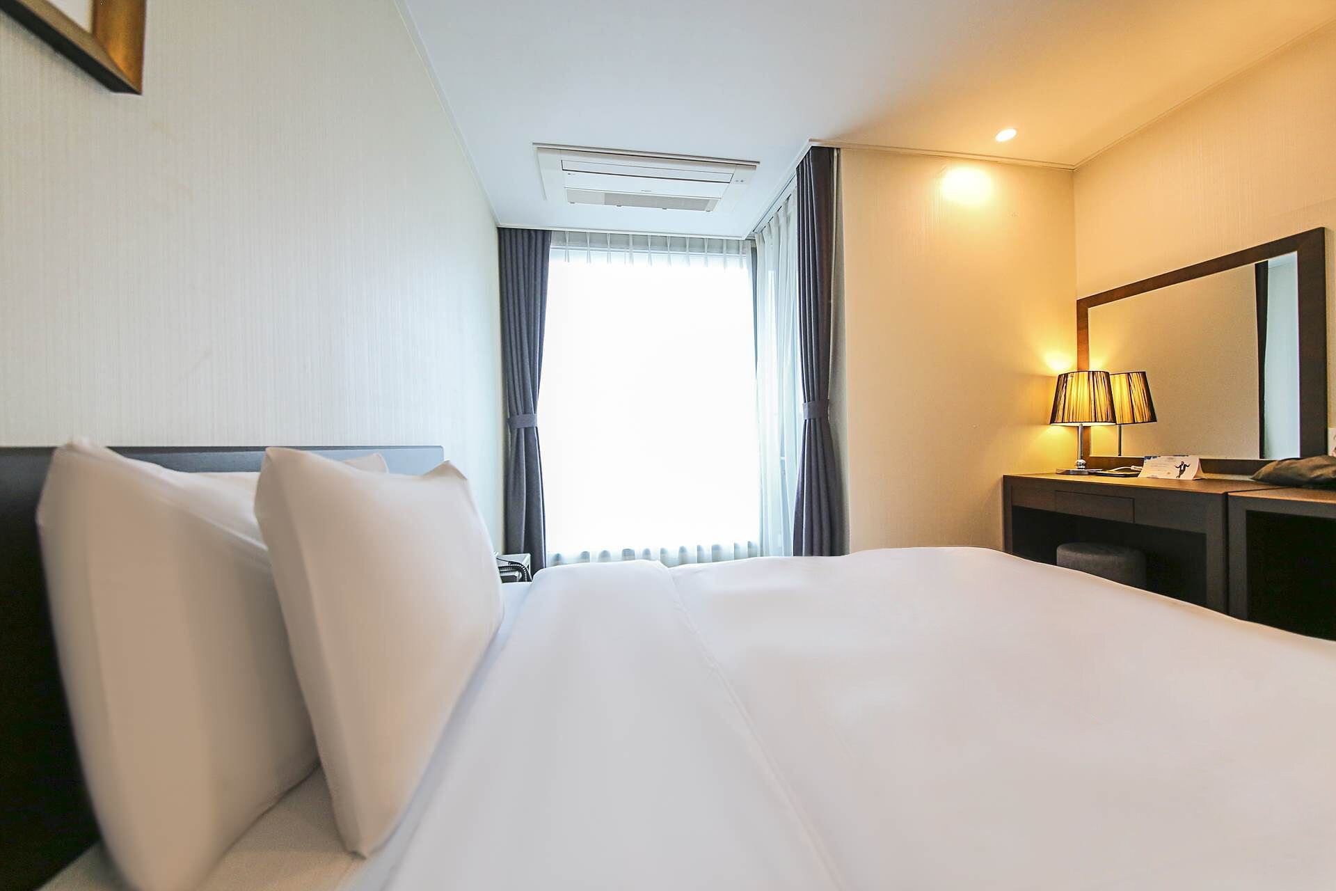 A hotel room with a bed, a desk with a lamp, a mirror, and a window with curtains.