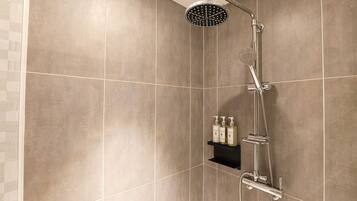A shower with a handheld showerhead and a wall-mounted control panel, a shelf with toiletries, and a tiled wall.
