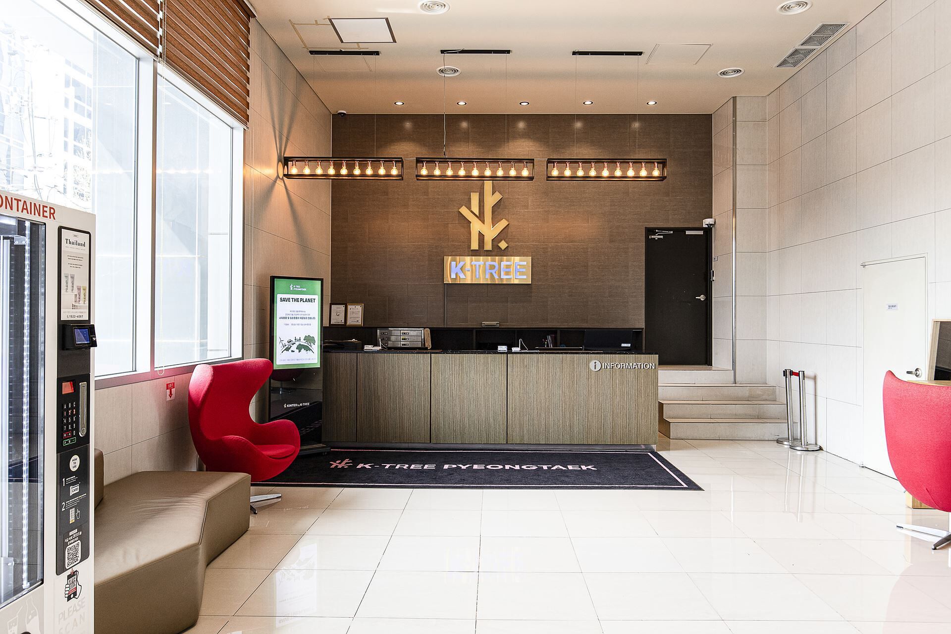 Una zona d'arribada moderna amb un taulell de recepció de fusta, un cartell que diu 'SAVE THE PLANET' i un senyal 'K-TREE'.