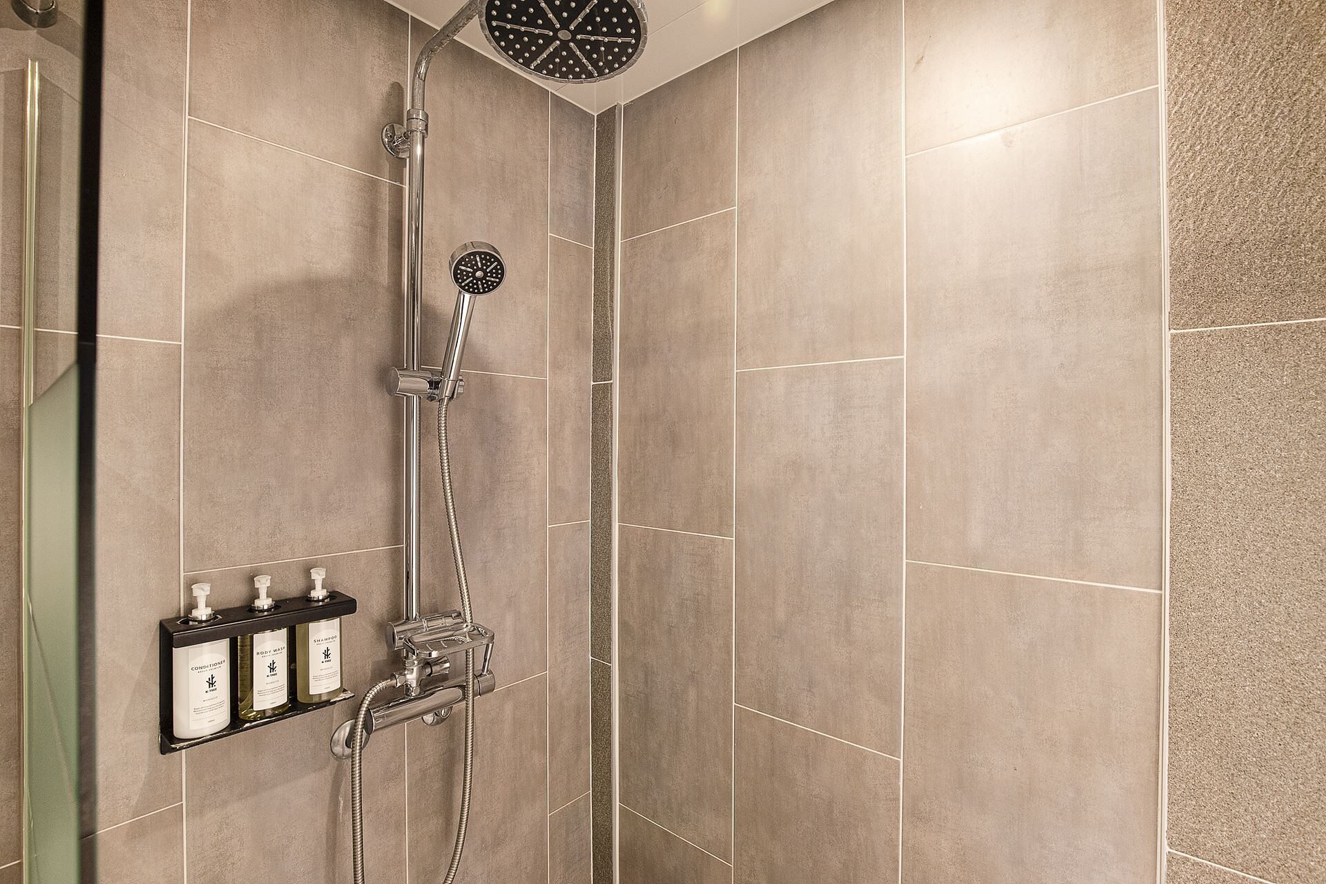 Doccia con soffione a mano e rubinetto a parete, ripiano con articoli da toilette e parete rivestita in piastrelle.