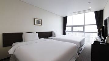 A hotel room with two beds, a large window, a framed picture on the wall, and a desk with a lamp.