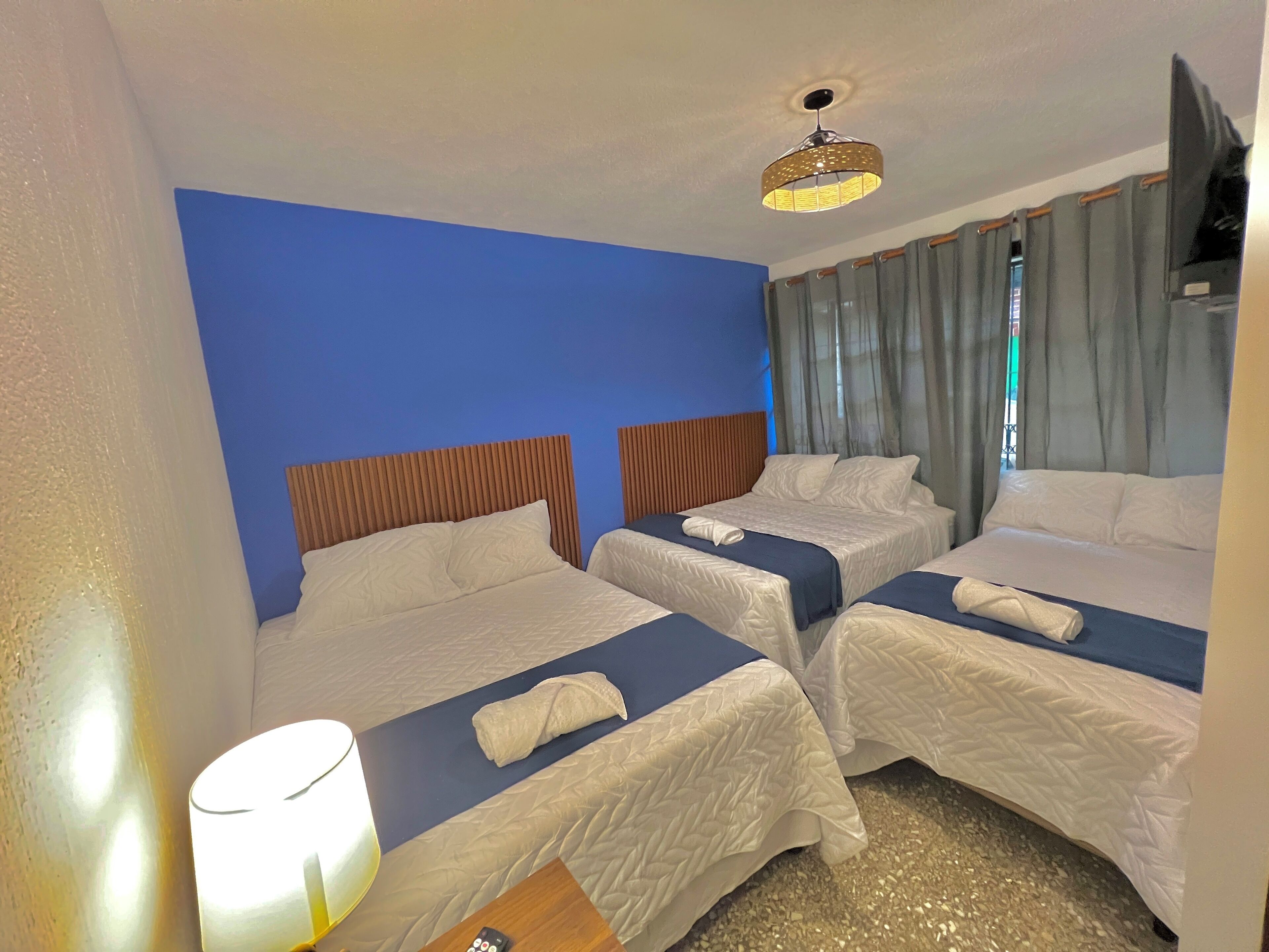 A hotel room with two beds, a blue accent wall, a ceiling light, and a window with curtains.
