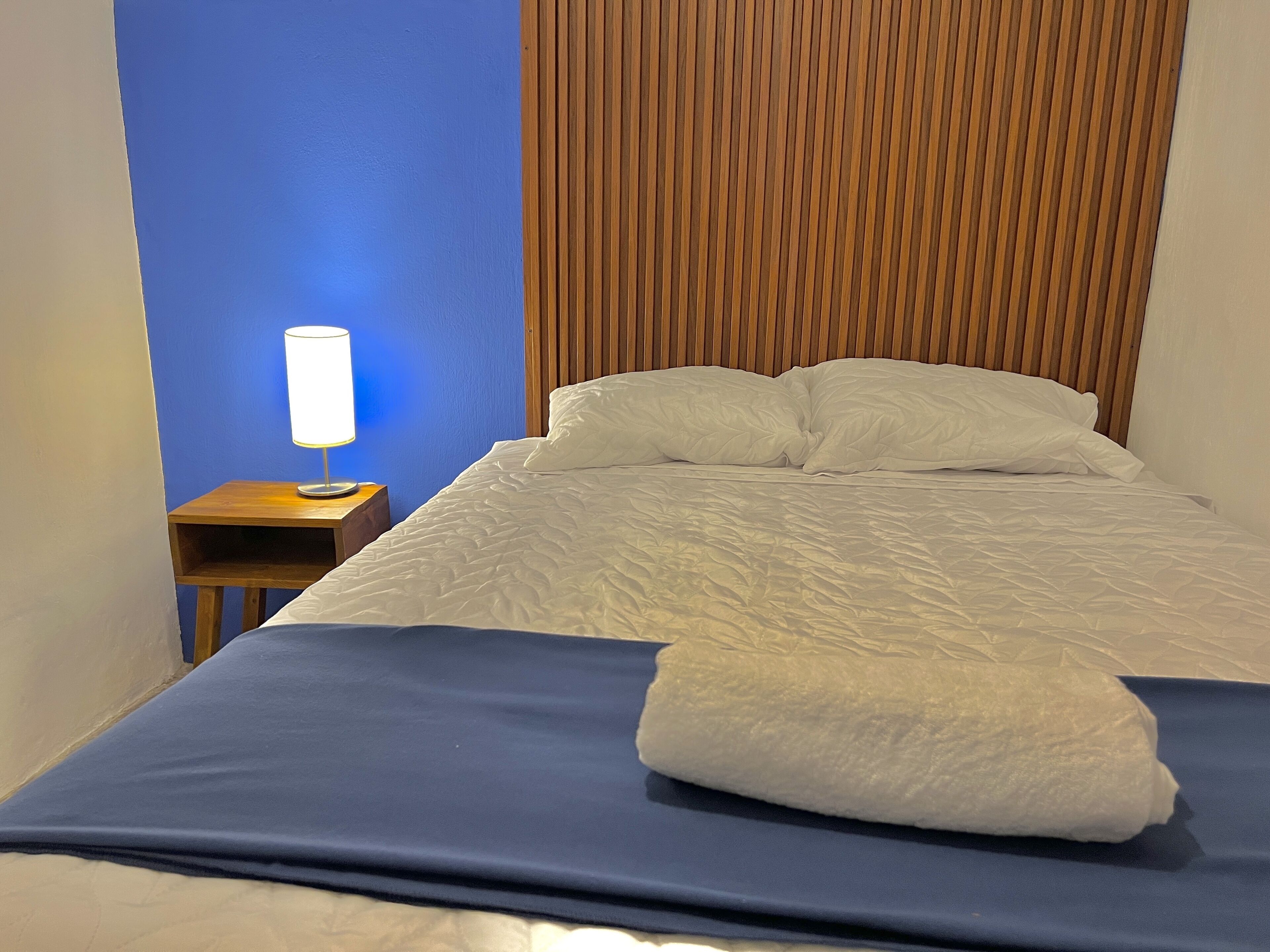 A neatly made bed with white linens, a blue blanket, and a folded white towel on the bed.