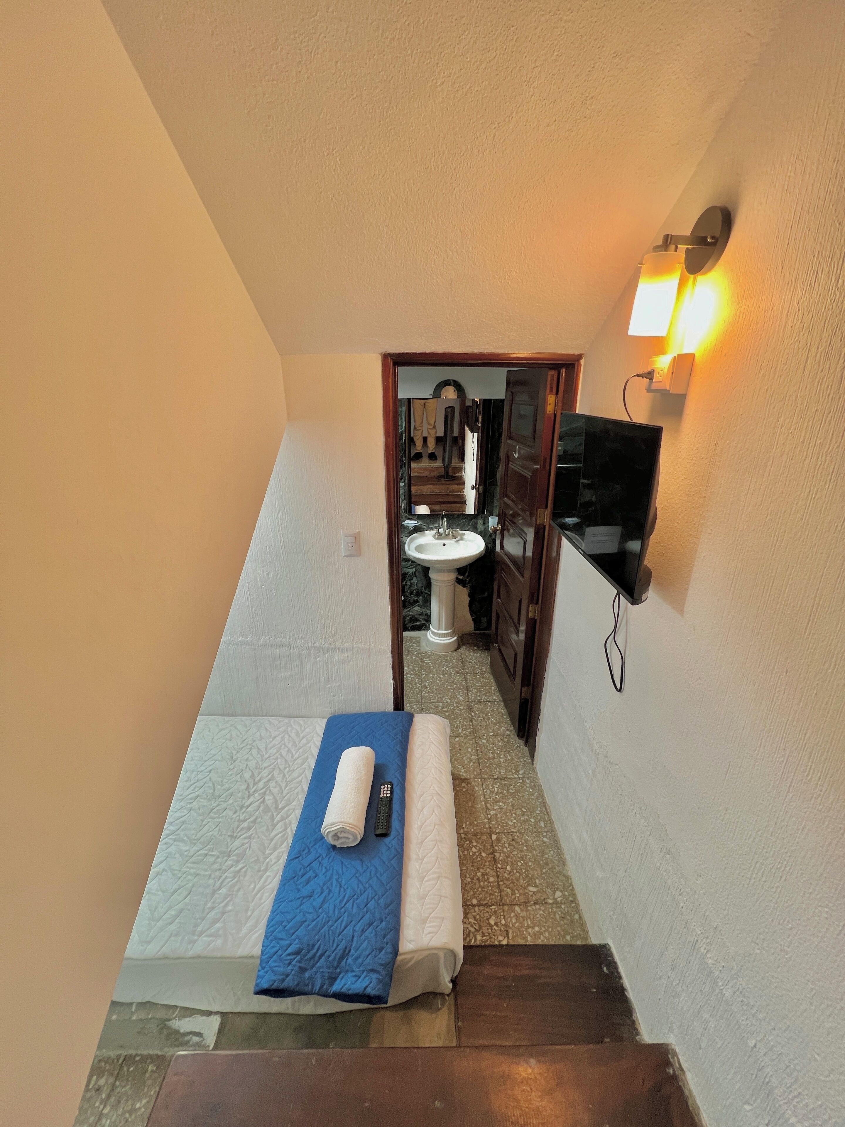 Un escalier étroit mène à une salle de bain équipée d’un lavabo et d’un miroir. On aperçoit un lit avec une couverture bleue et une télécommande sur la petite plateforme située au sommet de l’escalier.