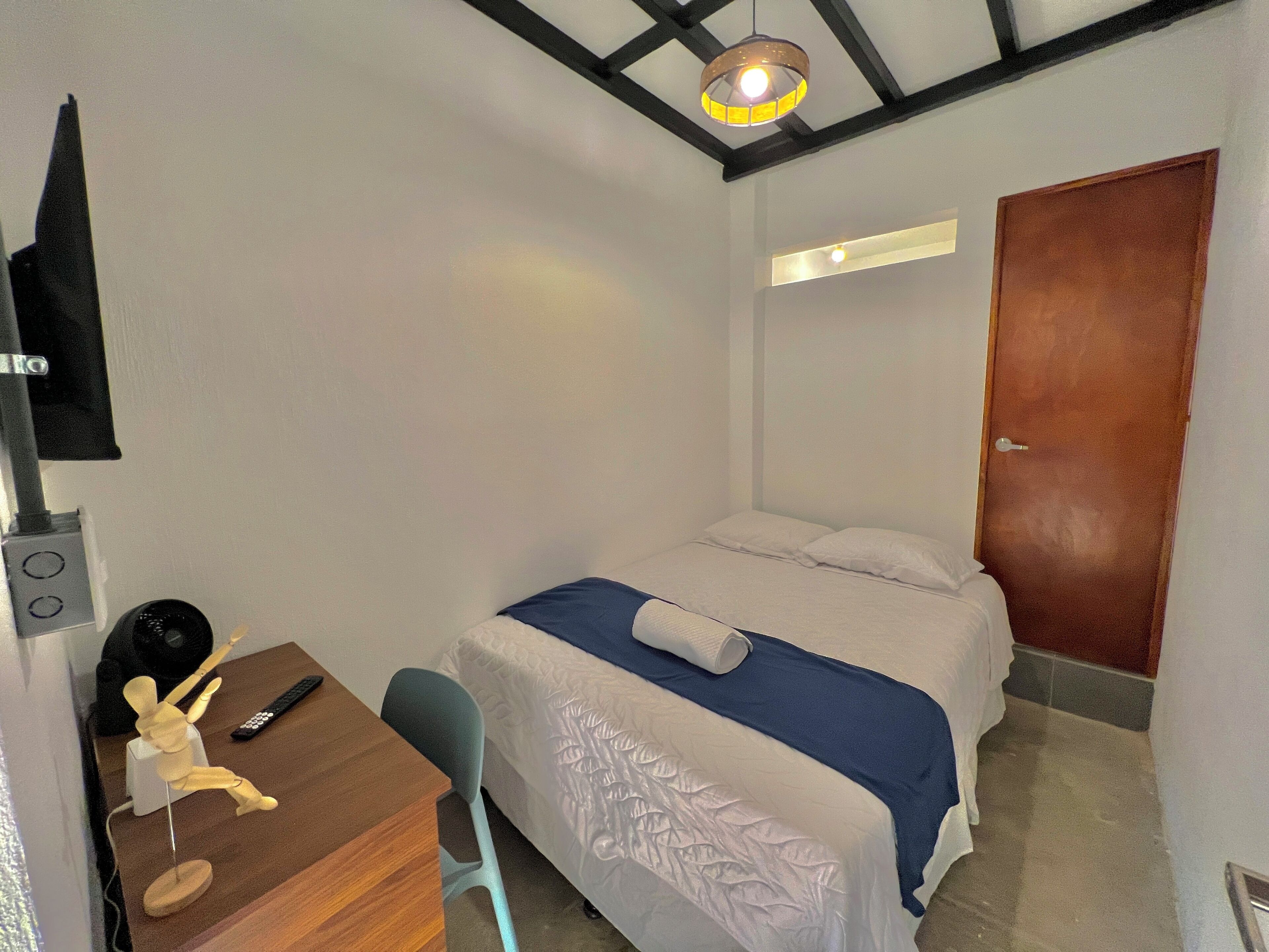 A small, neatly arranged bedroom with a bed, a desk with a chair, a wooden mannequin, and a television mounted on the wall.
