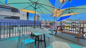 Un área de estar al aire libre con mesas y sillas, protegida por grandes sombrillas, con vista a una calle con edificios y una estructura de techo azul.