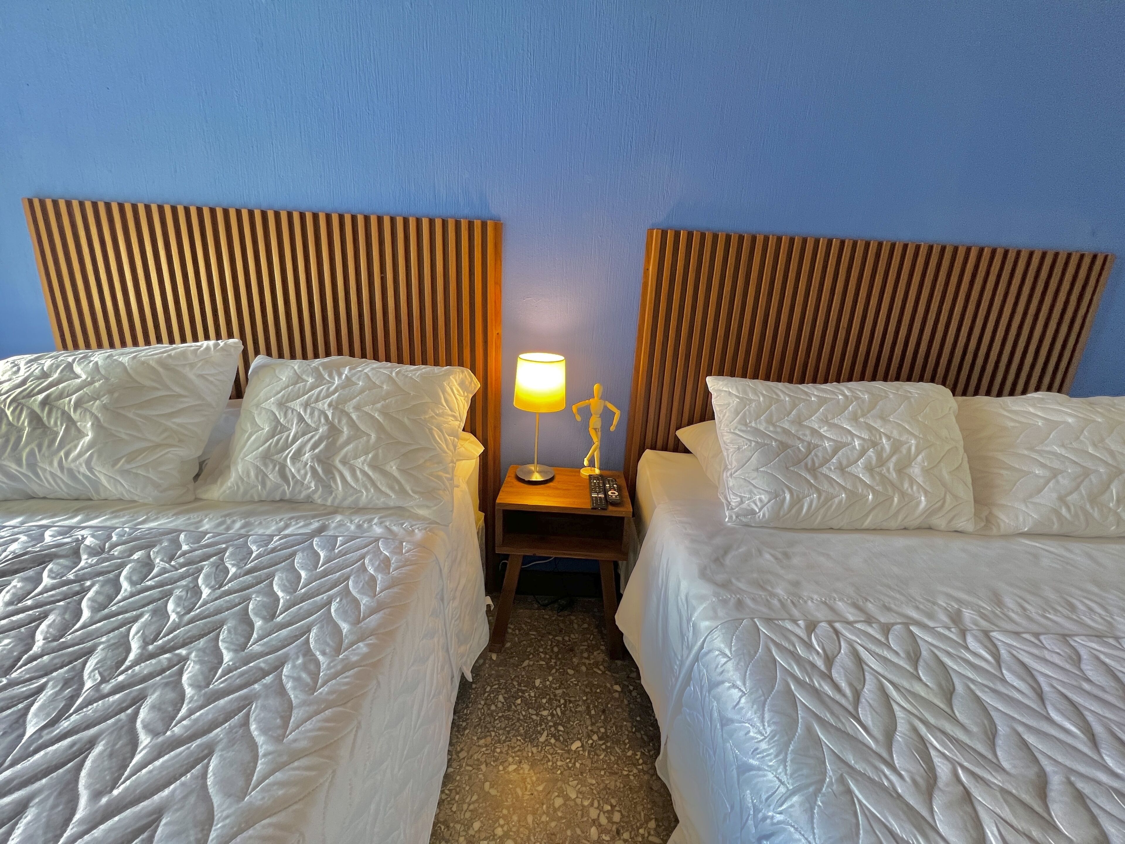 Habitación de hotel con dos camas, cada una con ropa blanca y un diseño acolchado. Entre las camas hay una mesita de noche con una lámpara y una pequeña estatua. La habitación tiene cabeceros de madera y una pared azul.