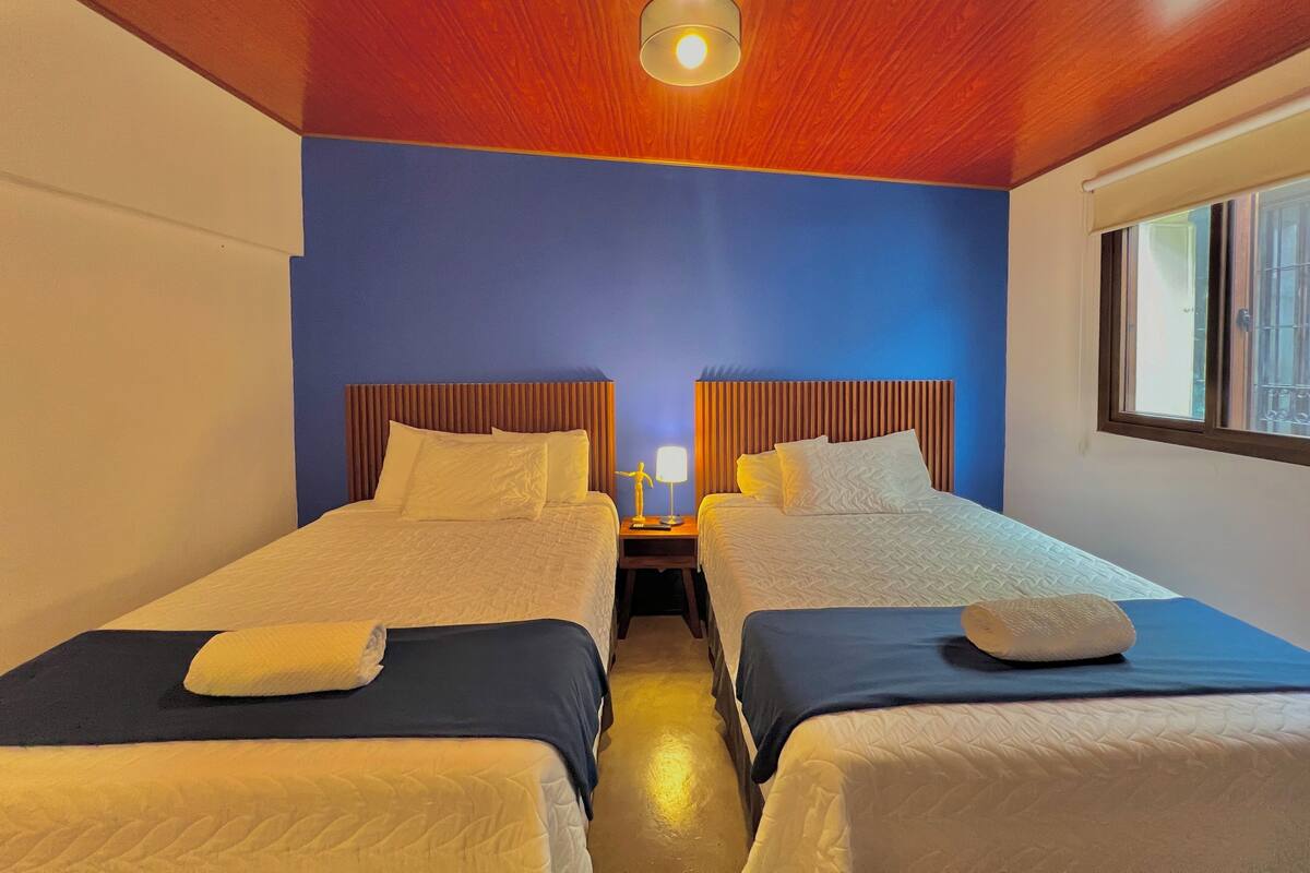 Une chambre d’hôtel avec deux lits, des têtes de lit en bois, une table de chevet avec une lampe et une fenêtre avec des barreaux.