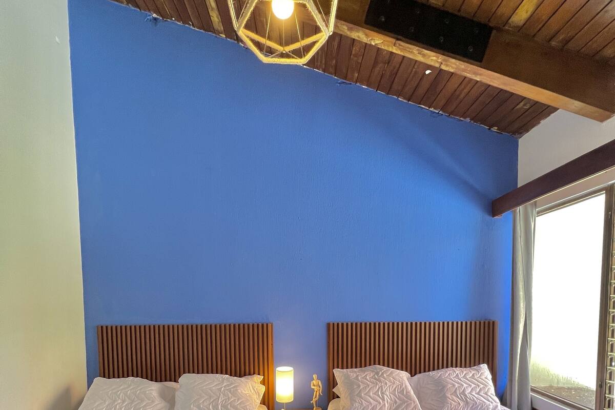 Une chambre d’hôtel avec deux lits, des têtes de lit en bois, un mur bleu, un luminaire suspendu et une fenêtre donnant sur un paysage.