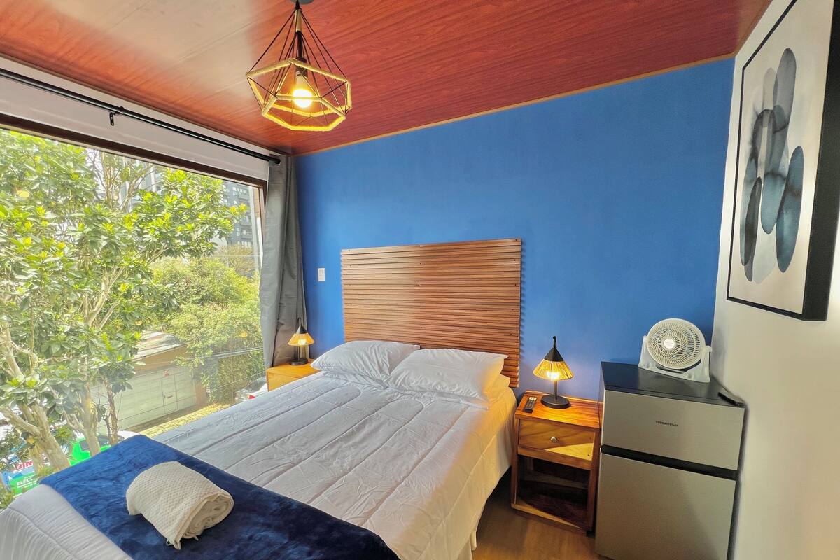 Une chambre à coucher avec un lit, des lampes de chevet, une table de nuit, un radiateur et une fenêtre donnant sur des arbres.