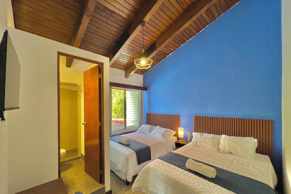 Une chambre d’hôtel avec deux lits, des têtes de lit en bois, un ventilateur de plafond et une vue sur l’extérieur.