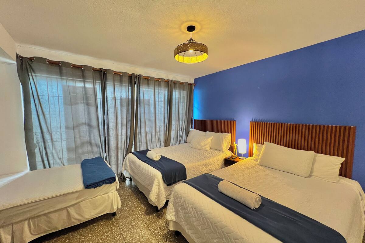 Une chambre d’hôtel avec deux lits, une grande fenêtre avec des rideaux, un luminaire au plafond et une table de chevet avec une lampe.