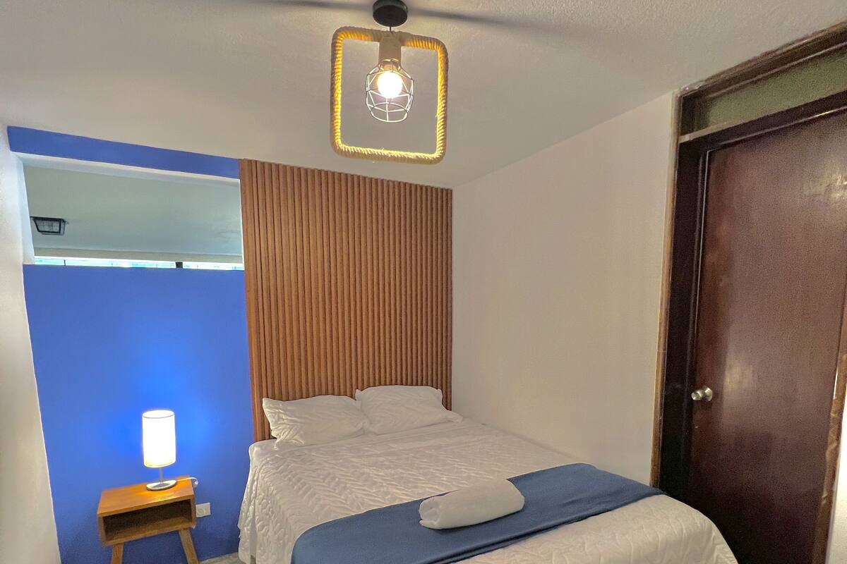 Une chambre d’hôtel avec un lit, une table de chevet avec une lampe, une tête de lit en bois, une porte et un luminaire au plafond.